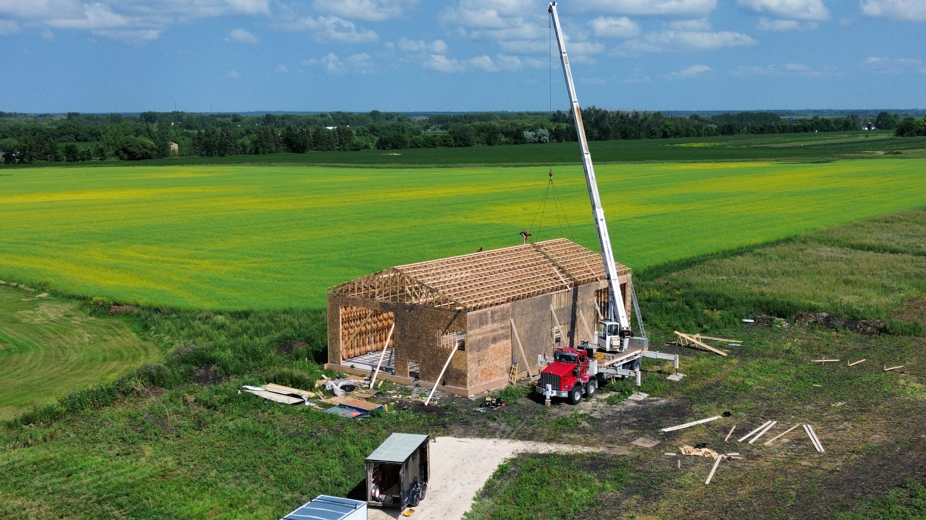 Rural shop build — aerial view, crane setting roof trusses over canola fields